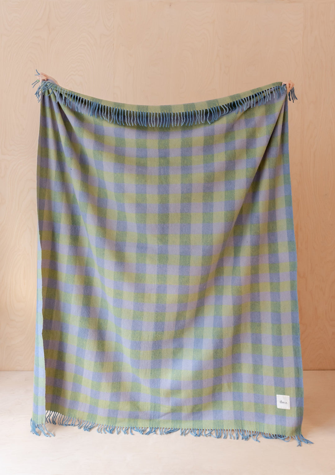 TBCo Recycled Wool Blanket in green and blue gingham pattern with fringe, displayed flat against wooden wall