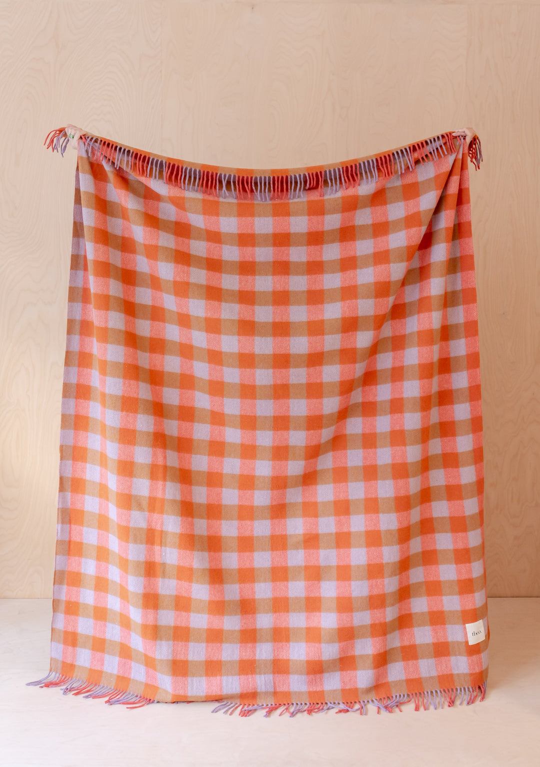 TBCo Recycled Wool Blanket in orange and lilac gingham pattern with fringe, displayed hanging against beige wall