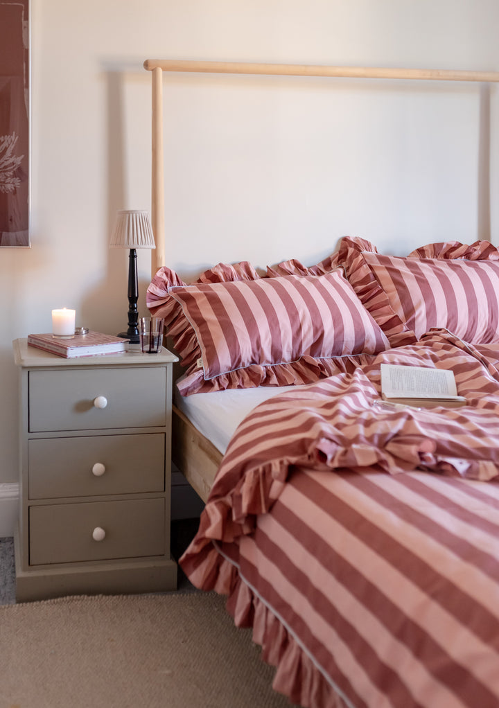 TBCo cotton pillowcases in pink and burgundy stripes with charming ruffled edges, styled on bed with grey nightstand