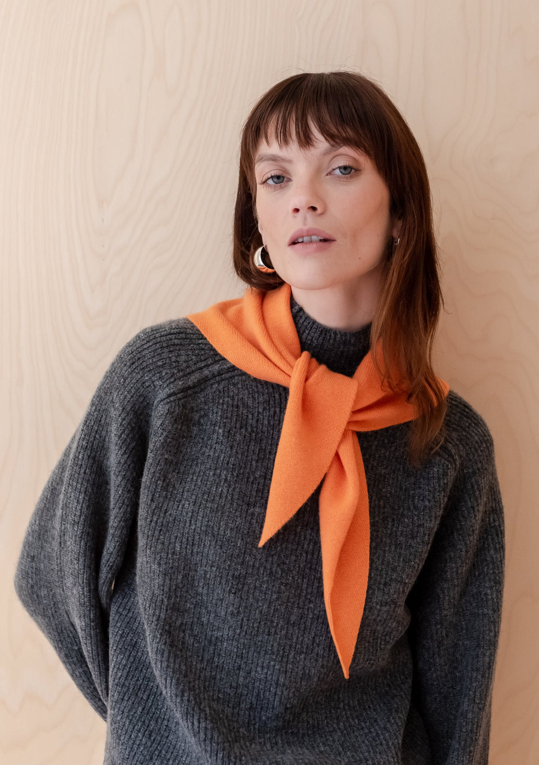 Merino Triangle Scarf in Apricot styled elegantly with a charcoal gray sweater against a light wooden background, showcasing the vibrant apricot color and versatile draping potential of the merino wool accessory.